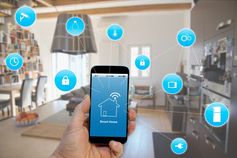 Hand hält ein Smartphone mit Icons zum Smarthome. Im Hintergrund ist ein Wohnzimmer zu sehen.