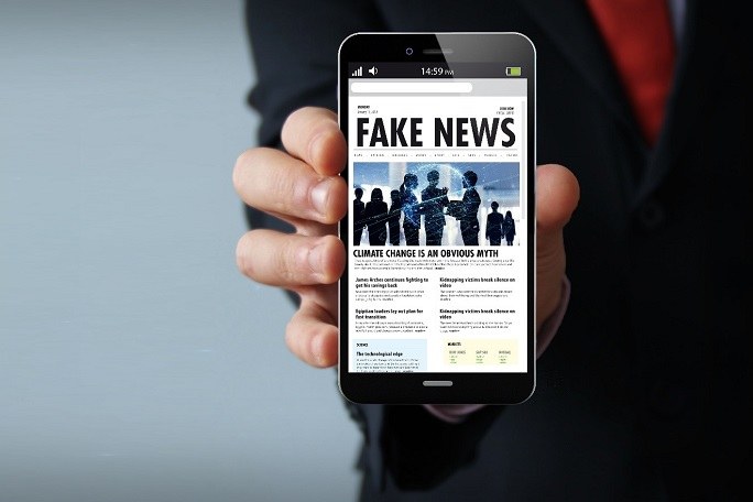 Eine Hand hält ein Smartphone mit dem Bildschirm nach vorne. Der Bildschirm zeigt eine Online-Zeitung mit dem Titel "Fake News"