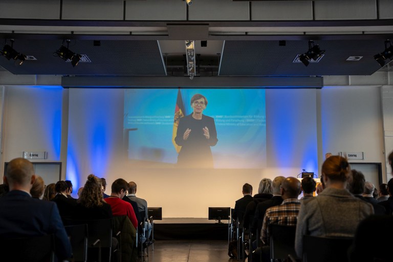 Menschen sitzen vor einer Bühne. Auf der Bühne ist eine Leinwand auf der Bundesforschungsministerin Bettina Stark-Watzinger zu sehen ist