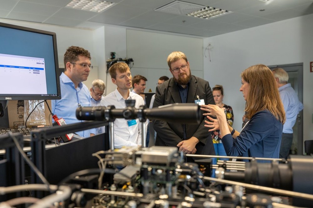 Kristian Döbrich, Toni Markurt (VDI/VDE-IT) und Daniel Behrendt (BMBF) im Austausch mit Marie-Theres Hahn (DLR-Institut für Kommunikation und Navigation) (v.l.n.r.). Im Vordergrund ist ein freistrahlbasiertes QKD-Übertragungssystem des DLR zu sehen. 